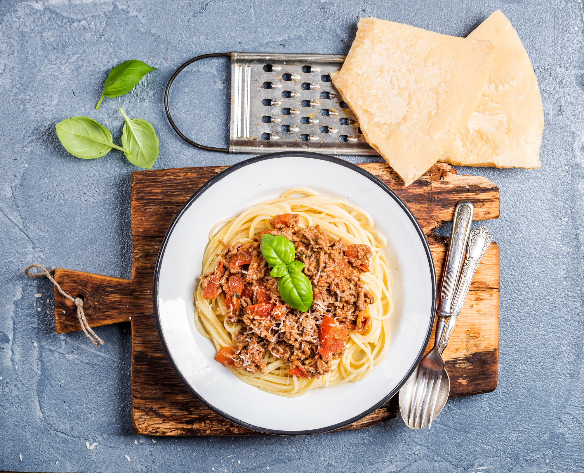 Pasta dinner. Spaghetti Bolognese in metal plate on rustic wooden board with Parmesan cheese, grater and fresh basil on grey concrete background, top view, horizontal